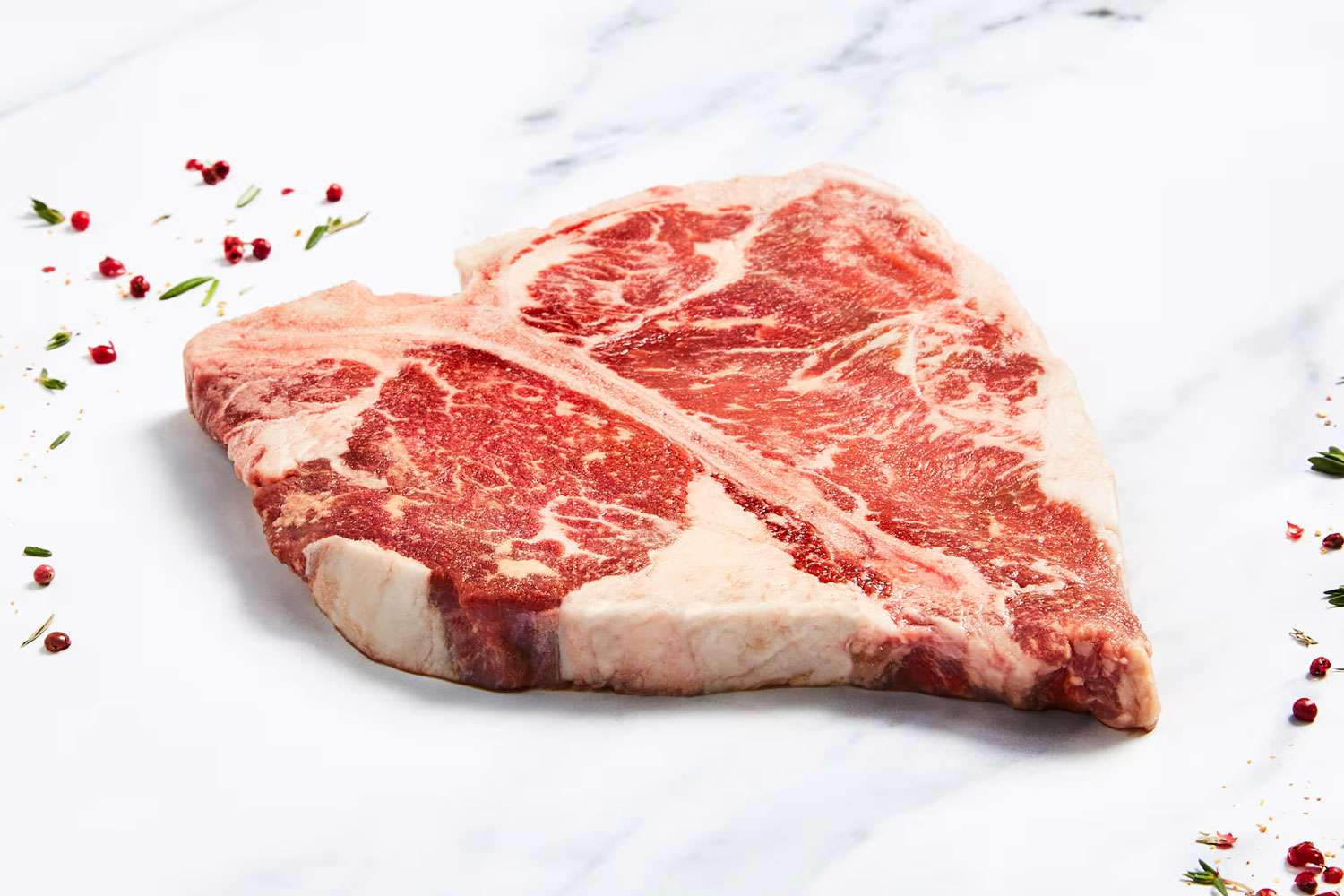 A raw Tbone steak on a white surface garnished with scattered herbs and peppercorns