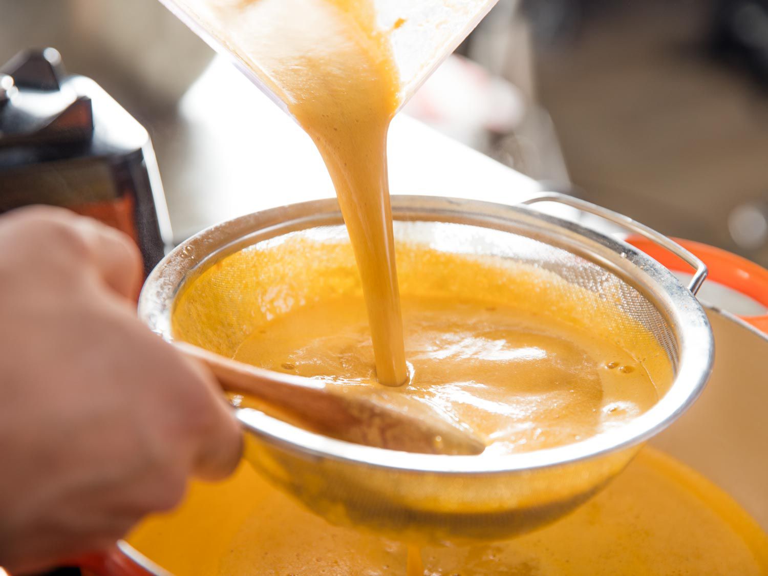Creamy, emulsified lobster broth being poured from a blender through a fine-mesh strainer, which is set over the Dutch oven.