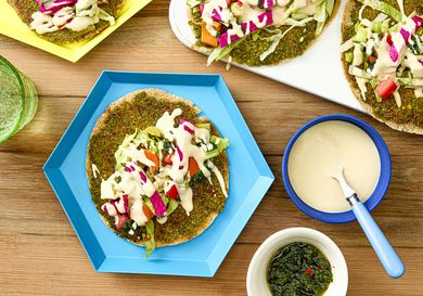 A smash falafel pita dressed with toppings next to more smash falafel pitas, a bowl of tahini sauce, and a bowl of zhug. 