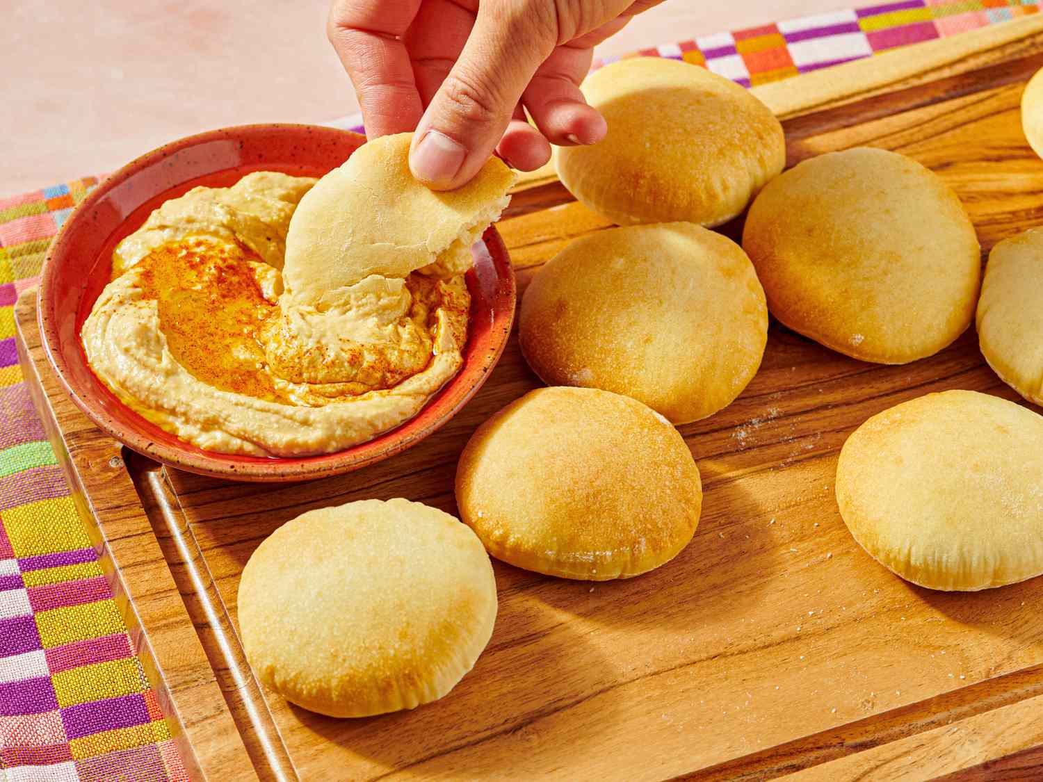 A hand dipping a piece of pita bread into hummus in a red bowl with several pita breads on a wooden tray nearby