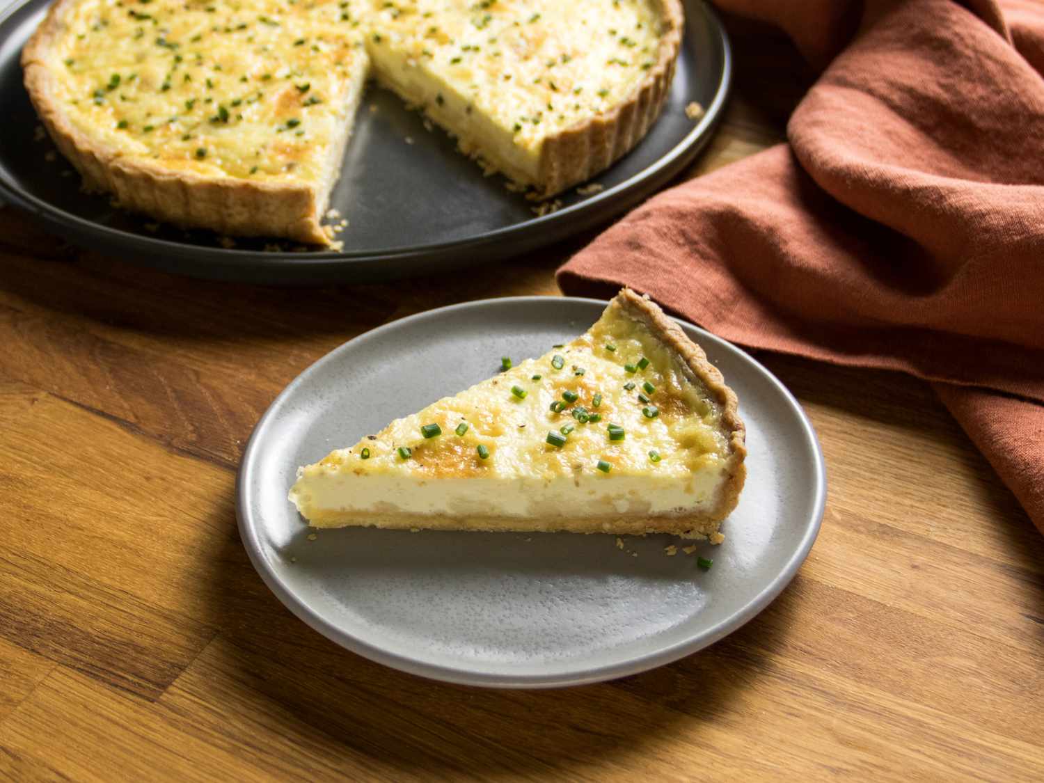 Slice of quiche on a grey plate with a brick colored textile to the side, and platter of rest of the quiche in the background