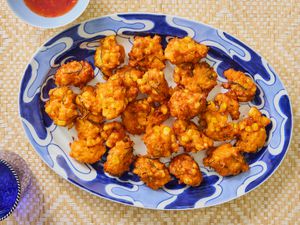 Fritters on a blue and white printed plate, on a bamboo mat. Glass of blue water, and red chili sauce on the side 