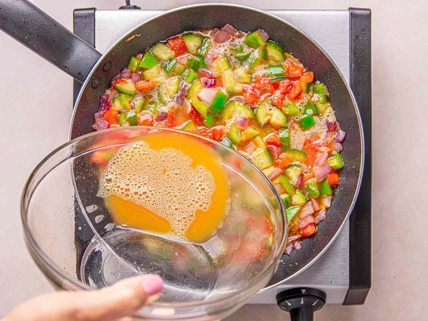A bowl of whisked eggs being poured into a pan filled with chopped vegetables on a stovetop