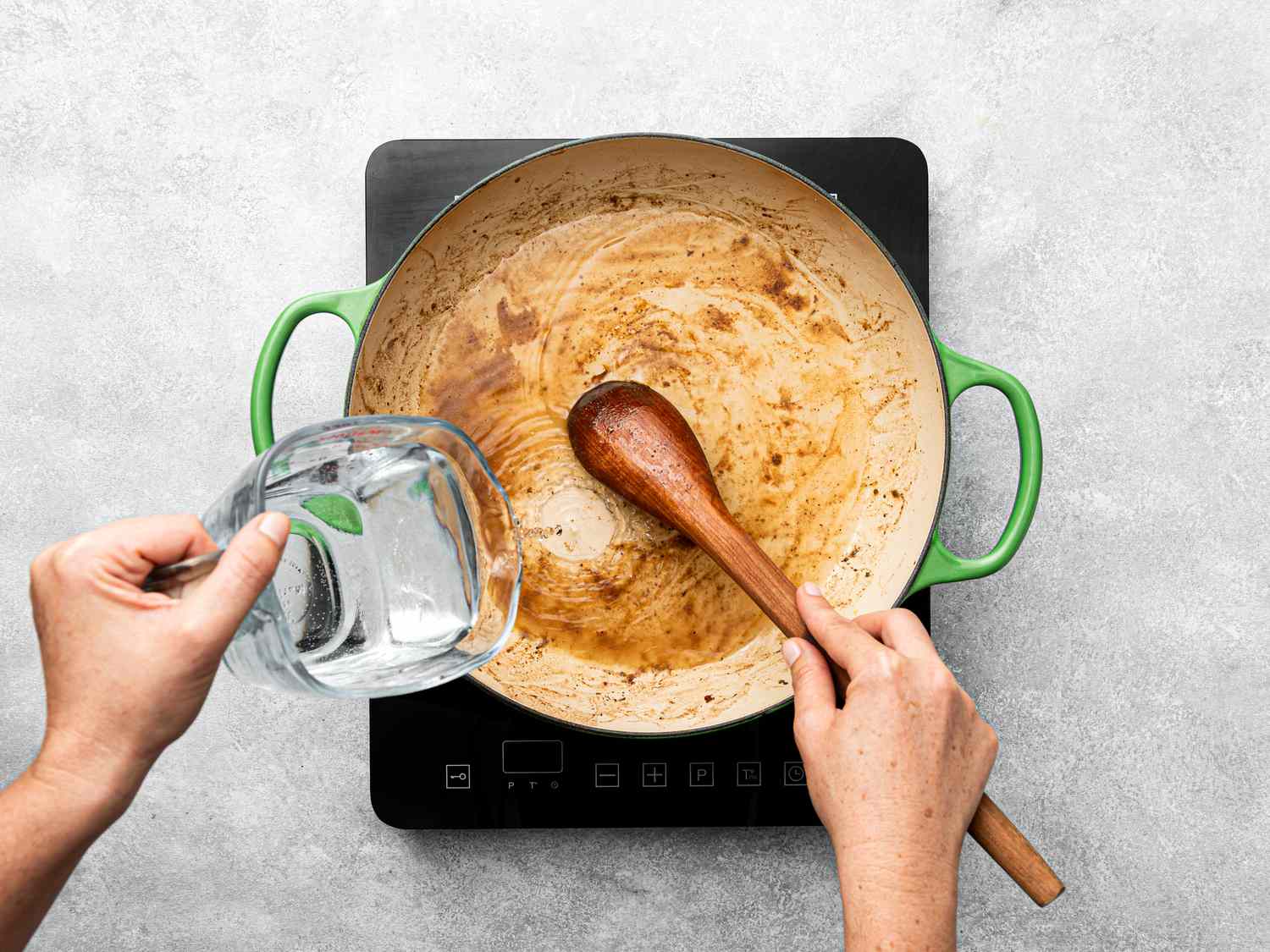 Hands deglazing a pan with water using a wooden spoon on a stovetop.