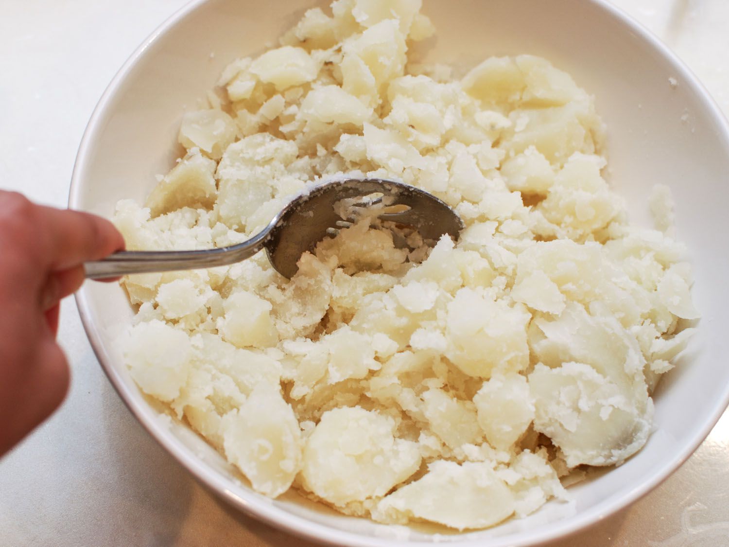 A bowl of cooked potatoes being broken apart and roughly mashed up into irregular chunks with a slotted spoon.