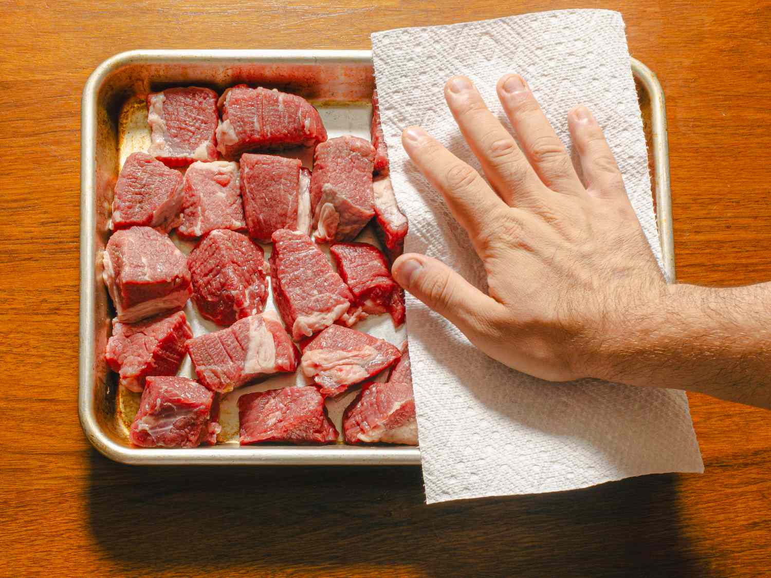 Chunks of beef being patted dry with a paper towel on a baking tray