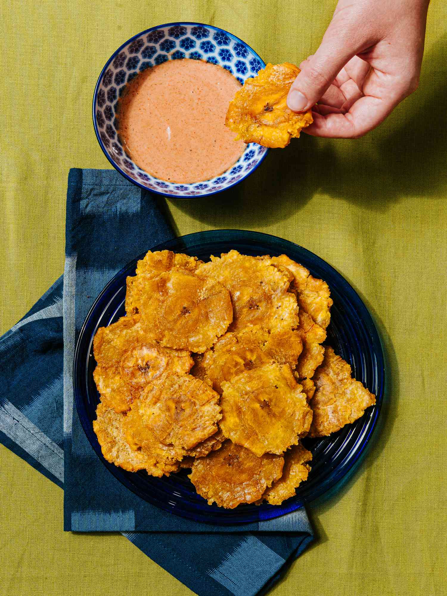 Overhead view of tostones on a blue plate with a blue napkin and a hand dipping a plantain in sauce