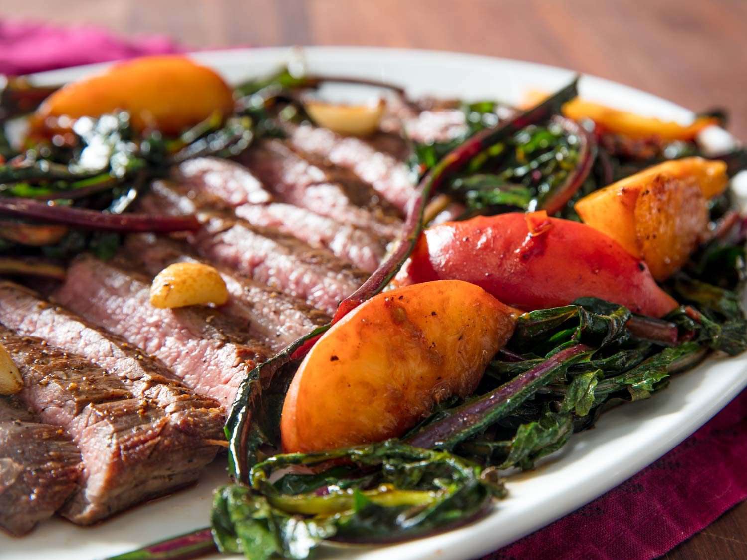 Close-up of sliced pan-seared flank steak accompanied by seared whole garlic cloves, seared peach slices, and wilted dandelion greens, on a white dish.