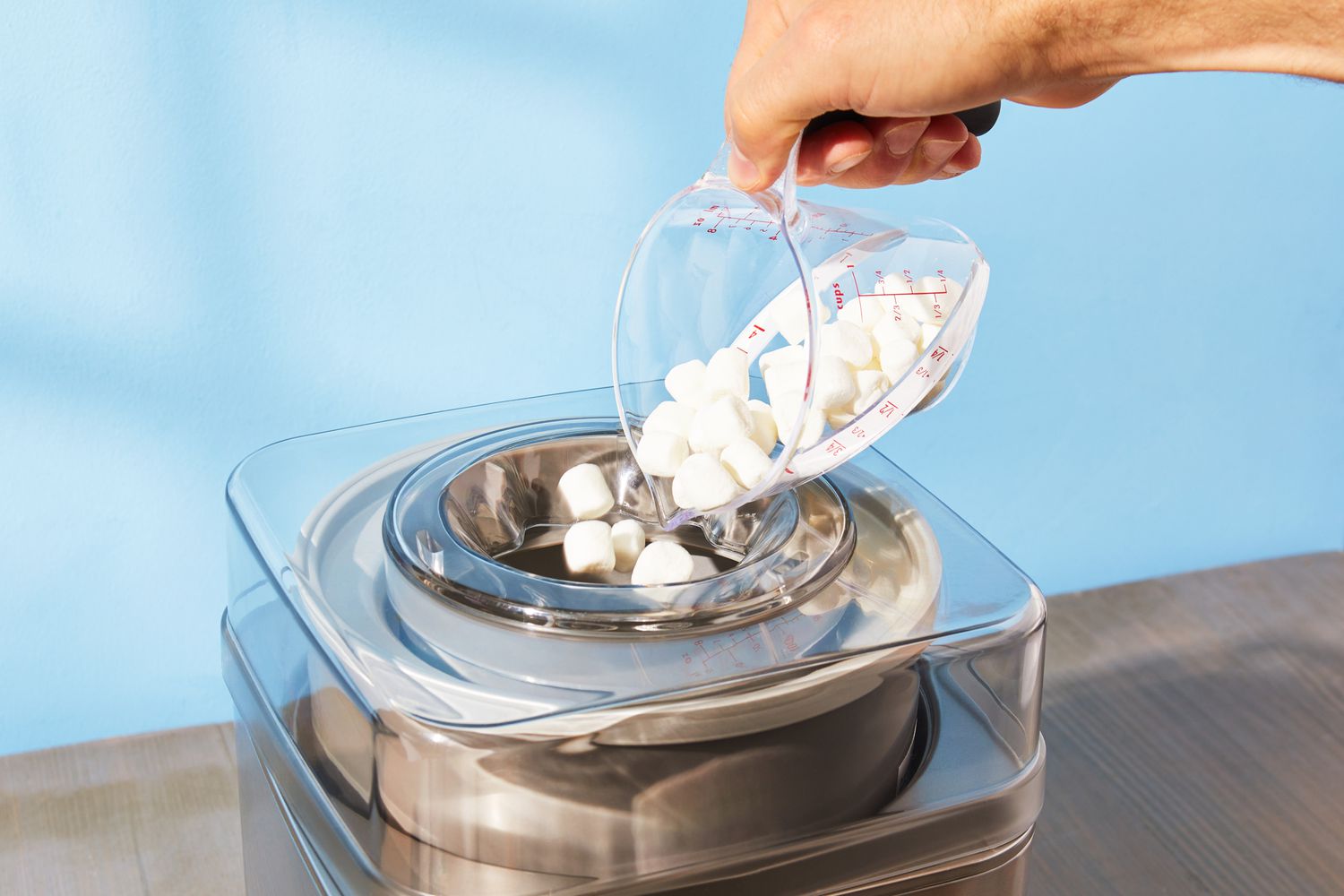 A person using a liquid measuring cup to add mini marshmallows through the top of the Cuisinart ice cream maker.