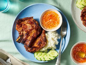 Pork chops over steamed rice white on a plate with sliced cucumbers and a ramekin of sauce