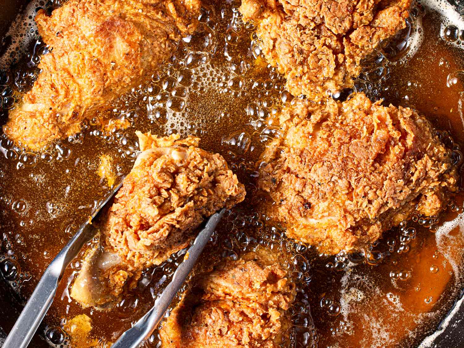 Closeup of fried chicken pieces being cooked in oil with tongs