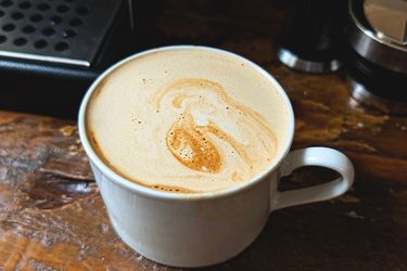 A cup of coffee on a wooden table with foam art on the surface, a coffee machine partially visible in the background