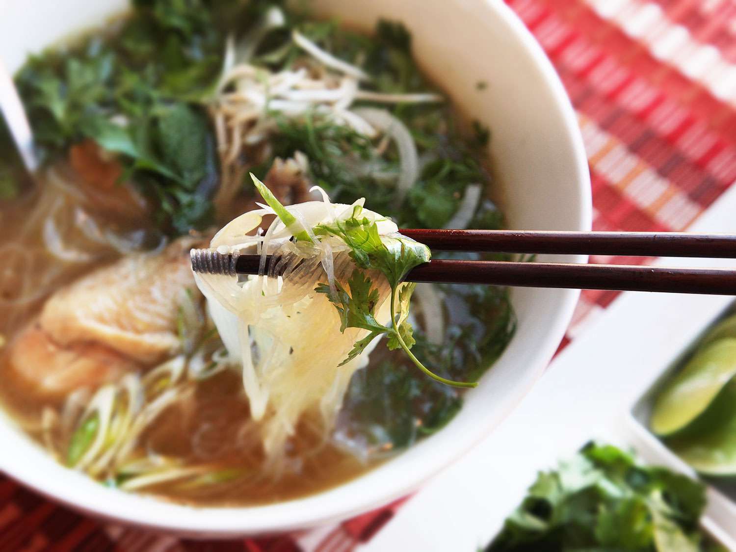 A bowl of pho ga. A pair of chopsticks are holding some noodles above the bowl. 