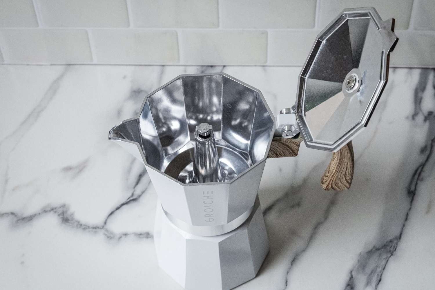 A stovetop espresso maker with its lid open on a marble surface