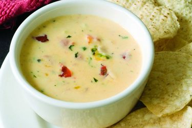 A small white bowl of chile con queso on a plate with some tortilla chips next to it.