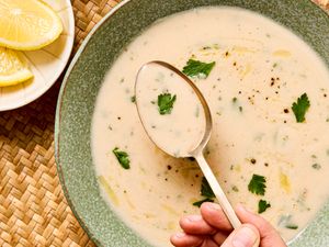 A bowl of garlic soup with garnish and a hand holding a spoon over the surface a plate of lemon slices to the side