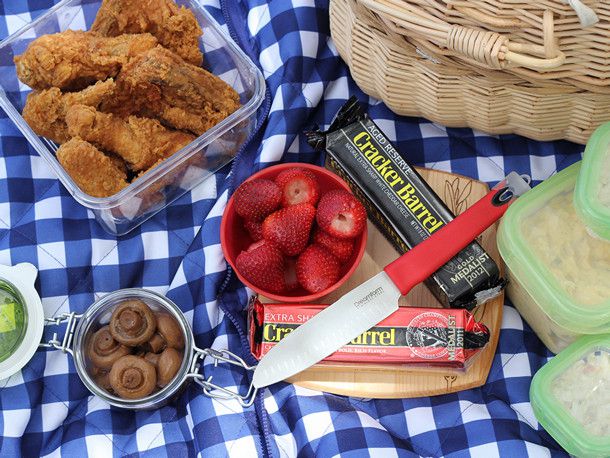 A blue and white checkered picnic blanket with a container of fried chicken, a picnic basket, containers of salads, two blocks of Cracker Barrel cheddar cheese, a container of strawberries and a container marinated mushrooms. There is a knife on the cutting board with the cheeses. 