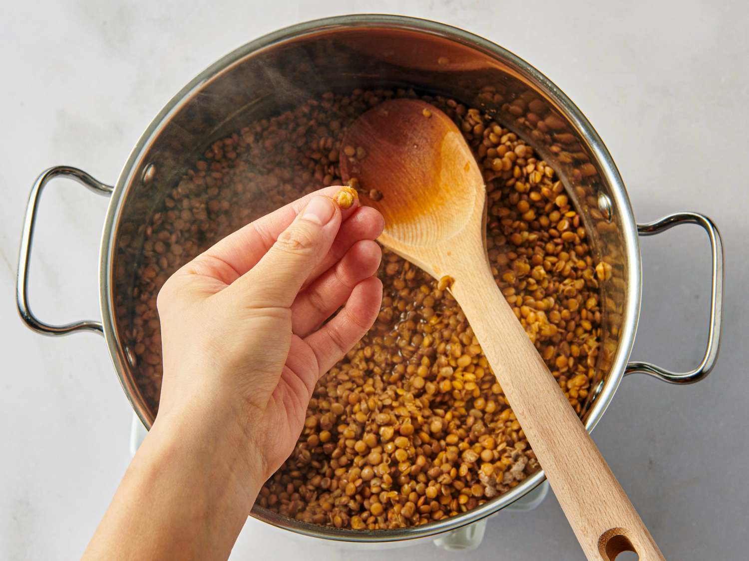 A pot of cooked lentils with a wooden spoon a hand holding a lentil above the pot