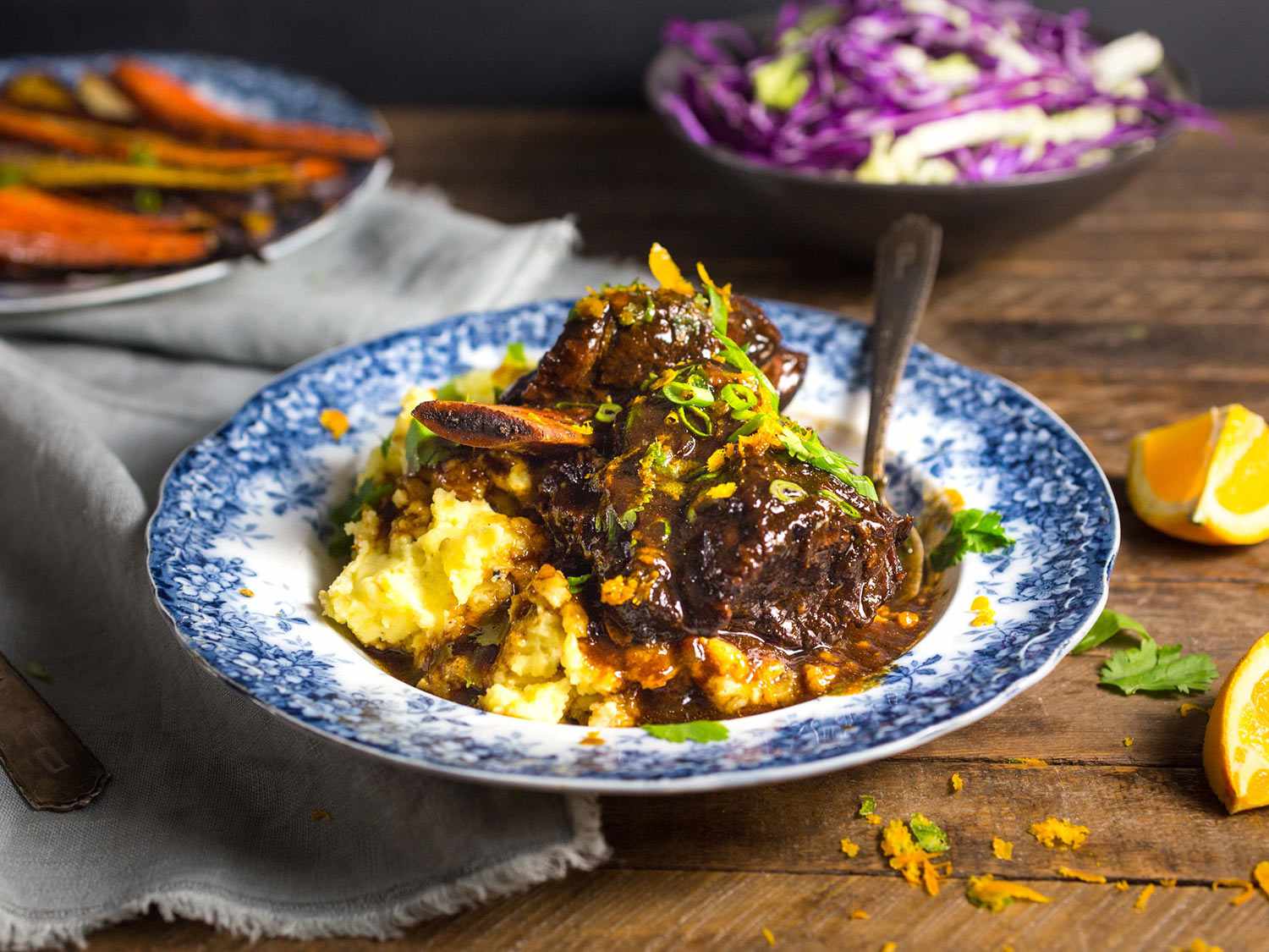 Blue and white bowl filled with braised shortribs and mash with bowls of cabbage and roasted carrots in background.