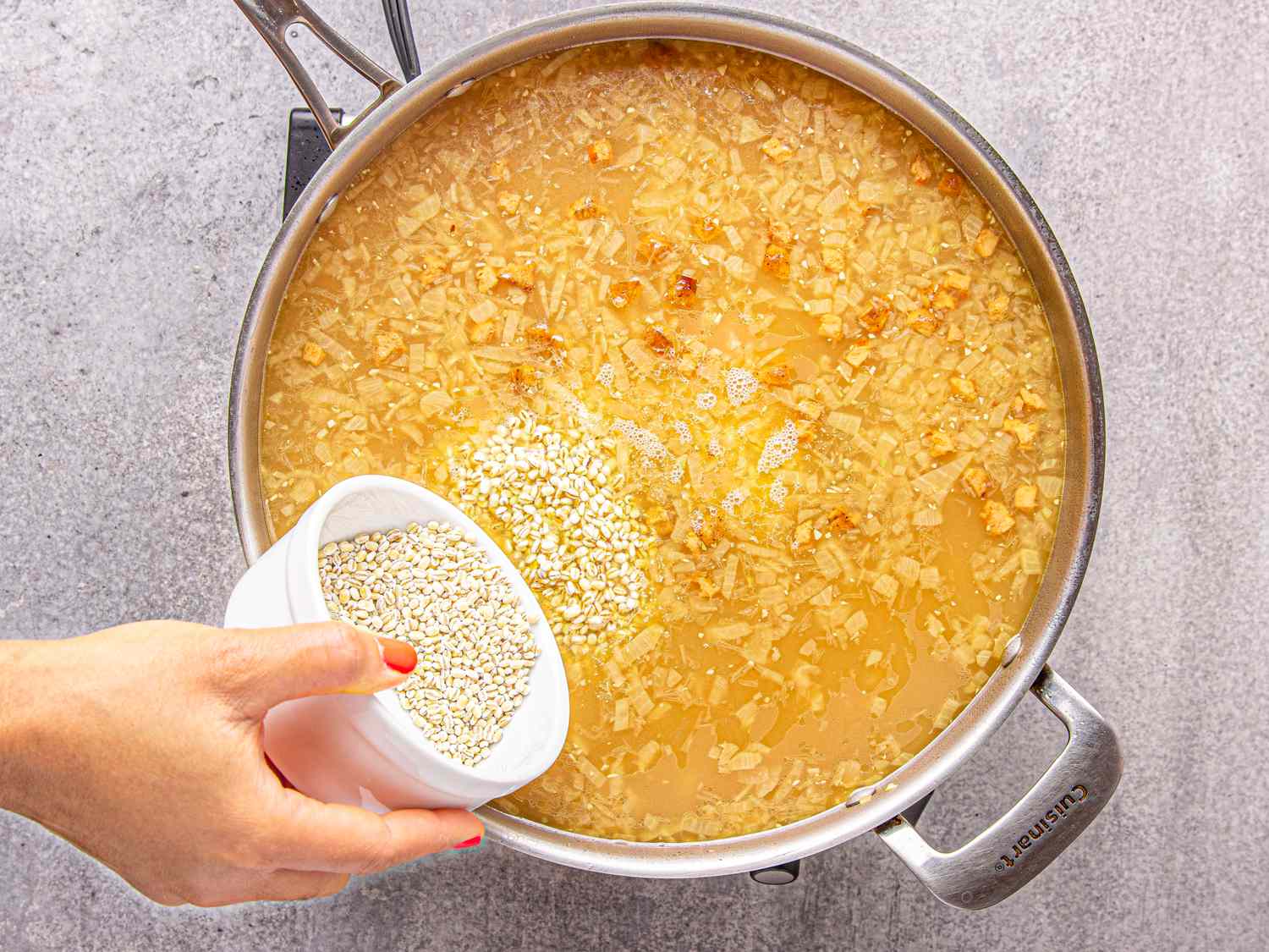 A hand adding a bowl of grains into a pot of soup on a stovetop
