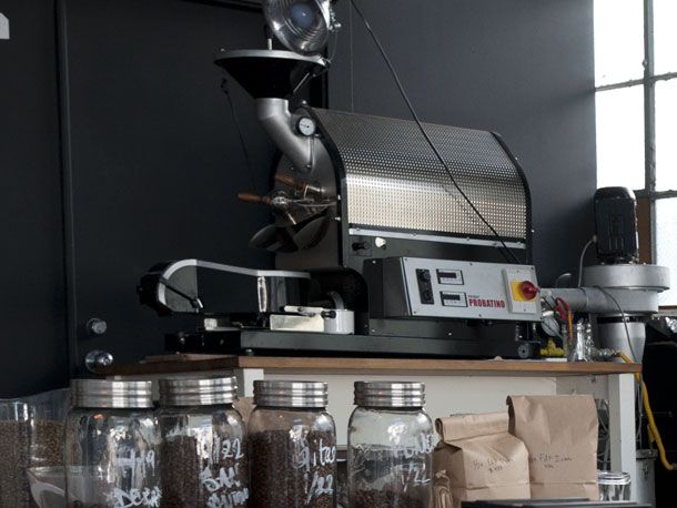 A photo of a small-batch coffee roasting machine on a counter. There are jars and paper bags of coffee grounds in the foreground of the image.