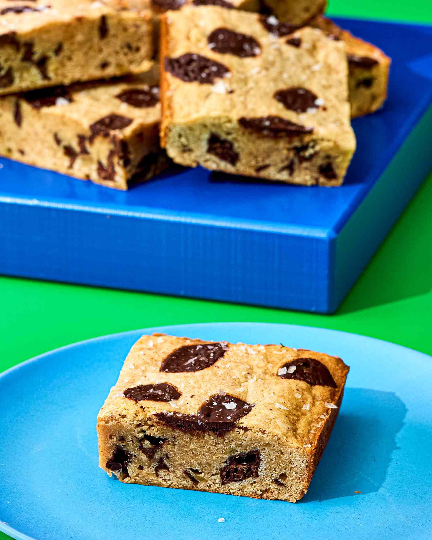 A piece of banana bread blondie with chocolate chunks on a blue plate additional pieces stacked on a blue tray in the background