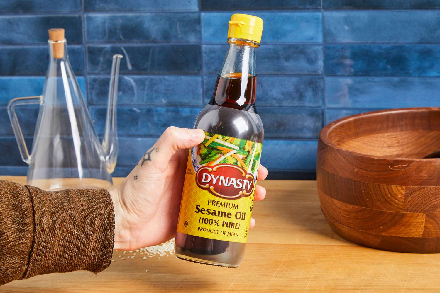 Hand holding a bottle of Dynasty premium sesame oil on a wooden counter, with kitchen items in the background