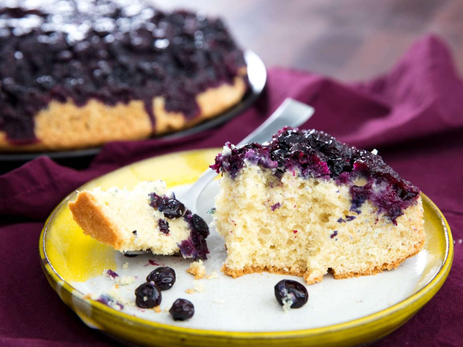 A plated slice of upside-down blueberry muffin. A bite-sized portion of the muffin is suspended over the plate on a fork.