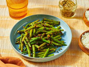 A plate of green beans seasoned with spices, next to a bowl of rice and a jar of seasonings on a textured surface