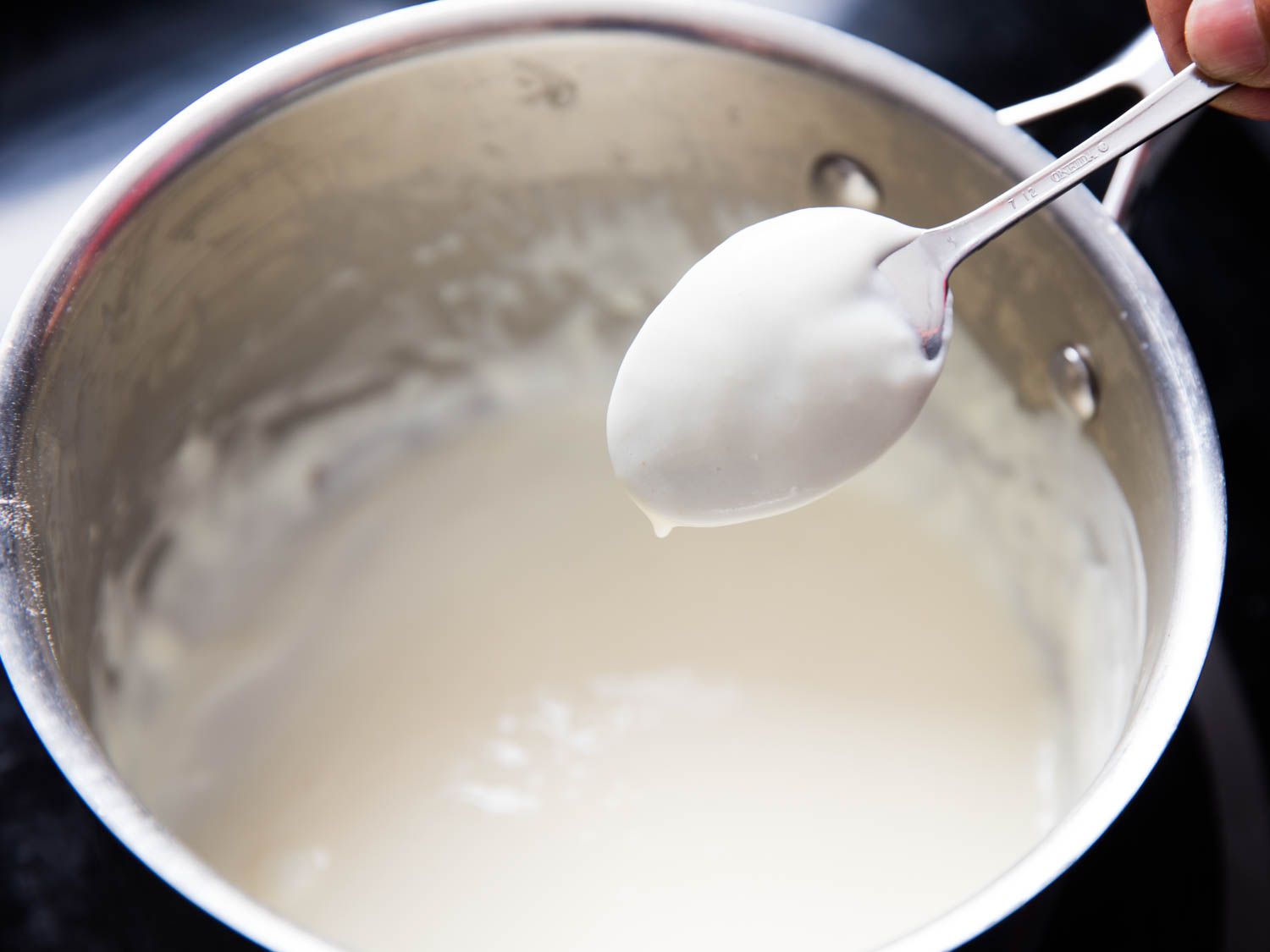 Author holding a dripping spoon coated with béchamel above a saucepan.