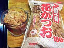 A glass measuring cup full of dried bonito flakes with a package of bonito flakes next to it.