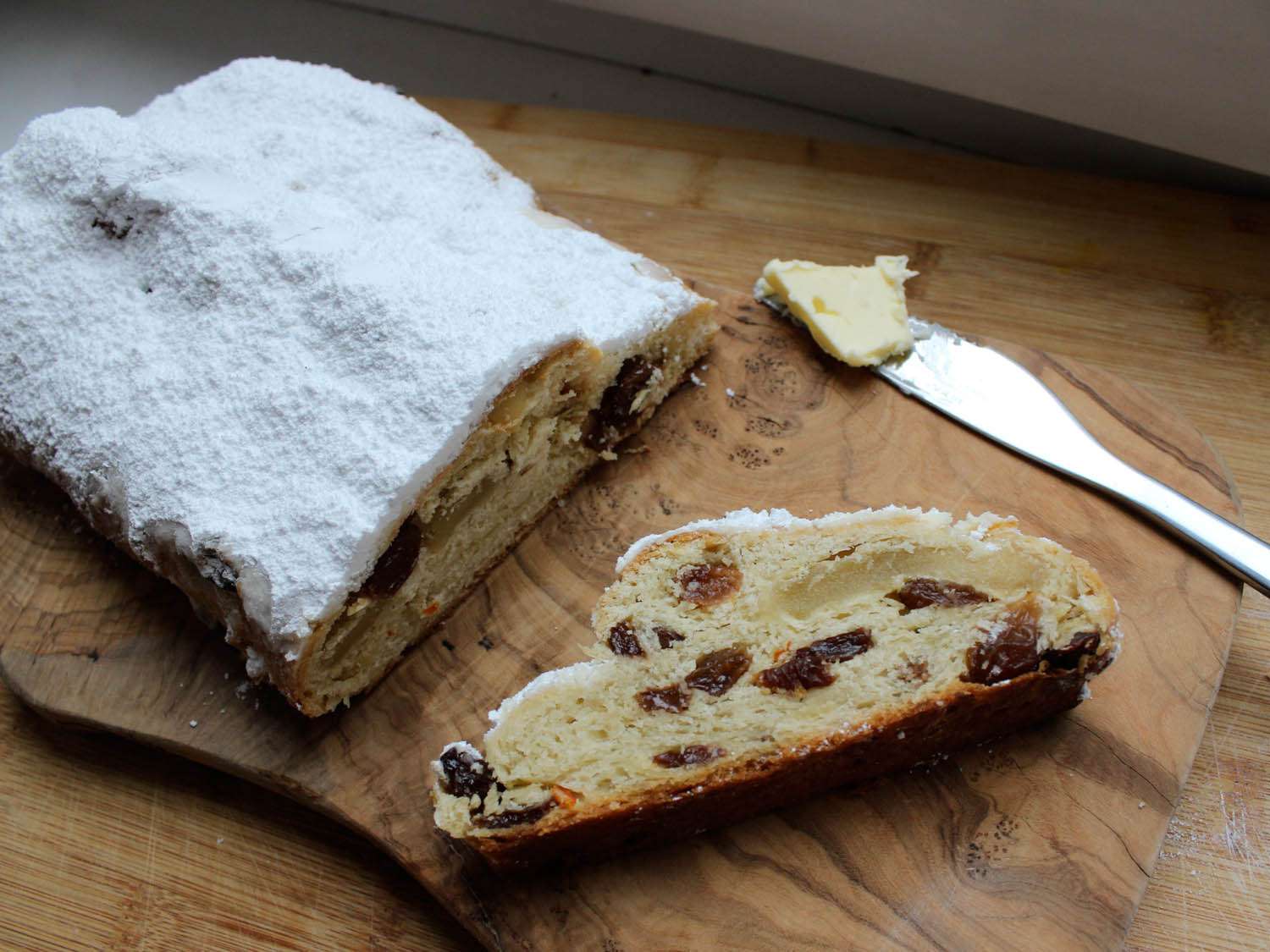 A finished loaf of stollen, studded with fruit, on a wooden cutting board next to a knife with butter on it.