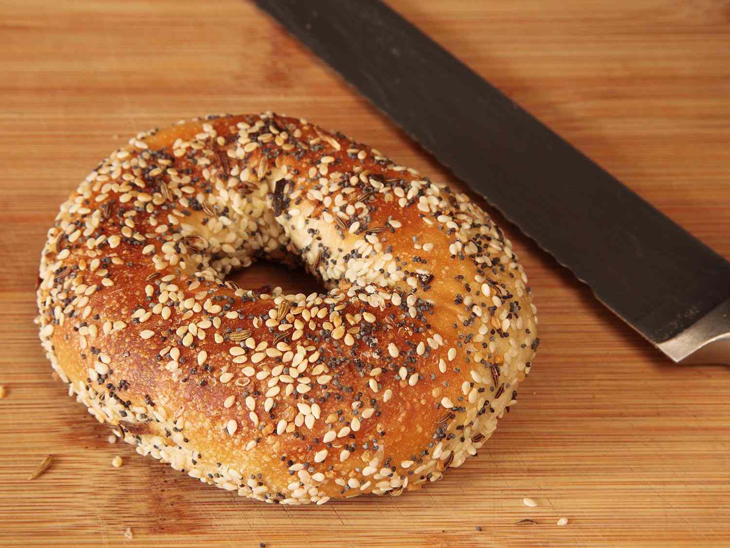 A whole everything bagel resting on a cutting board next to a knife