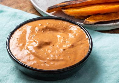 A bowl of chipotle mayonnaise on a light blue linen, with a plate of roasted vegetables behind it.