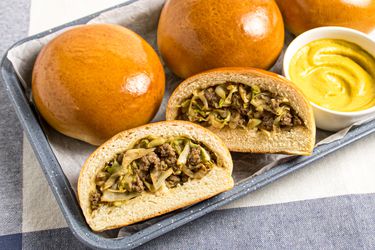 3 Bierocks on a platter with small white mustard dish, with a 4th bun cut open and filling is visible. Platter is on a grey white and blue striped textile. 