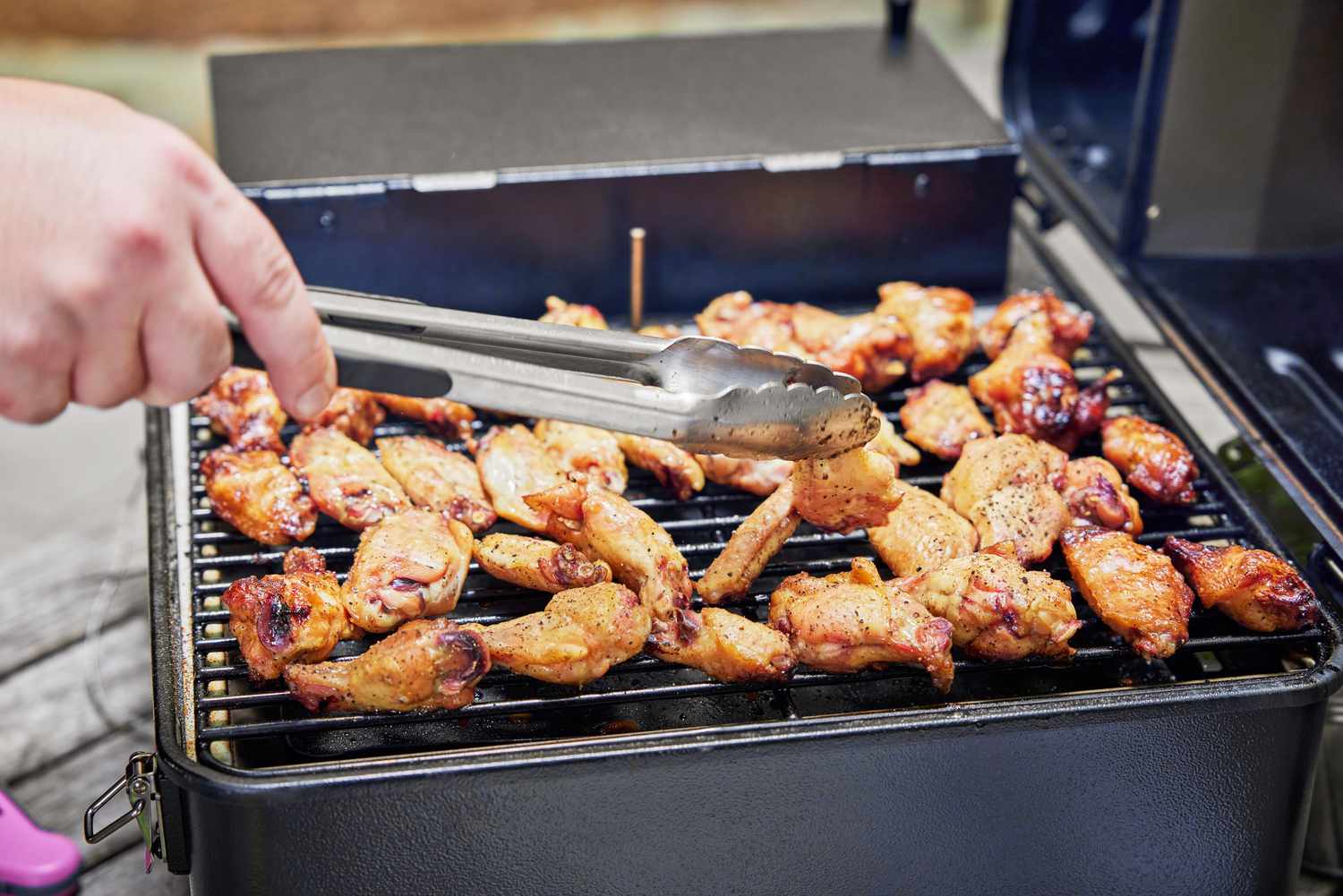 A hand holding tongs and flipping a chicken wing on a tabletop Traeger grill