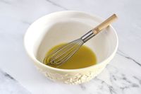 A whisk resting in a Mason Cash bowl containing a yellow liquid on a table surface