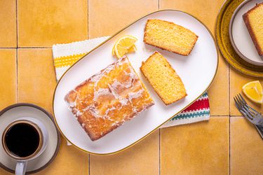 Platter with sliced lemon cake on yellow tile. On the side is a slice of cake, forks, and coffee mug
