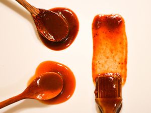Overhead image of two wooden spoons and a brush that have been doused in barbecue sauce and arranged on an off-white surface.