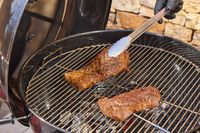 A hand using tongs to move steak on the SNS Grills Slow 'N Sear Original 22-Inch Kettle Grill