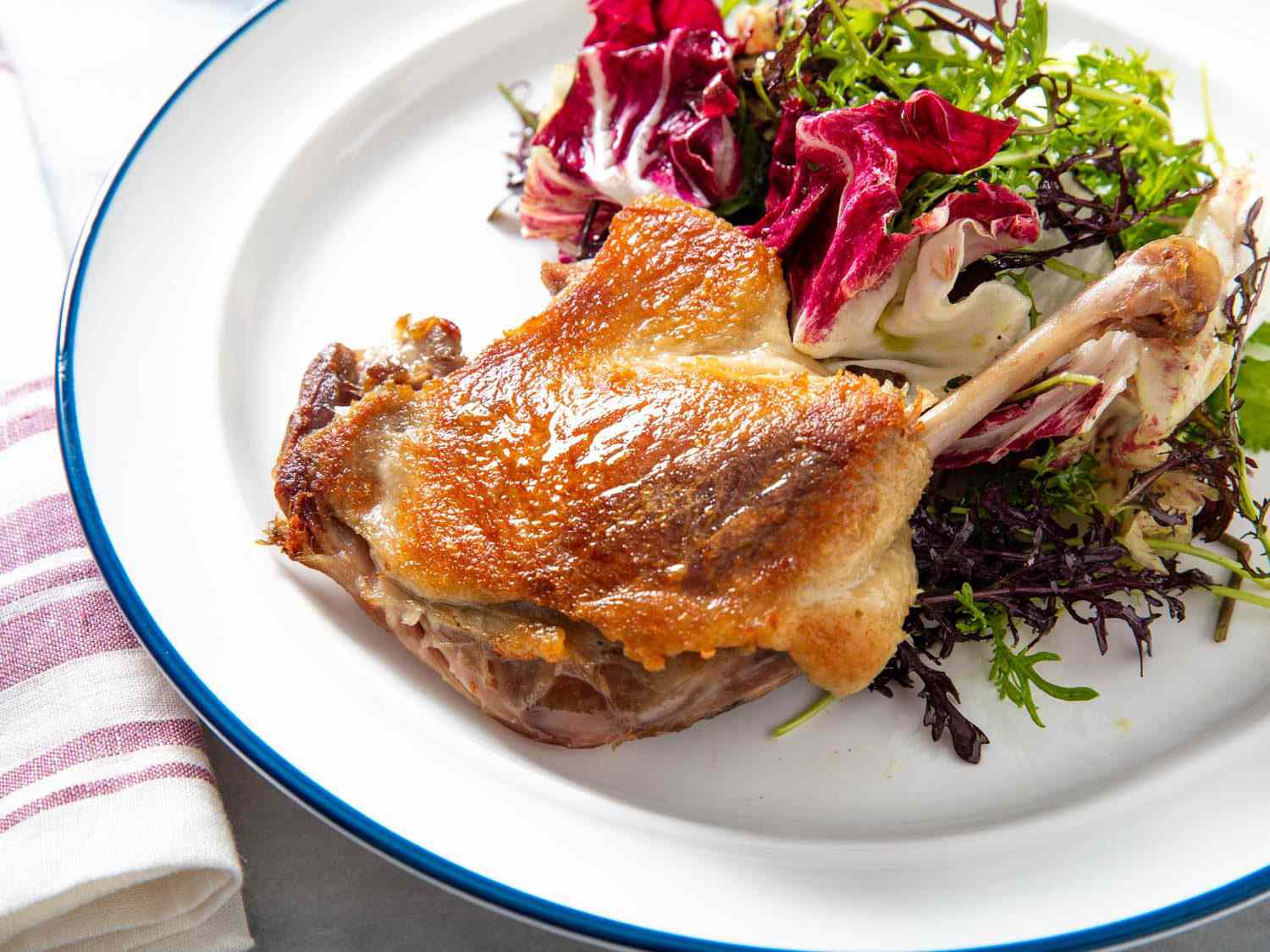 Closeup side view of a crisped confit duck leg with a mixed greens salad.