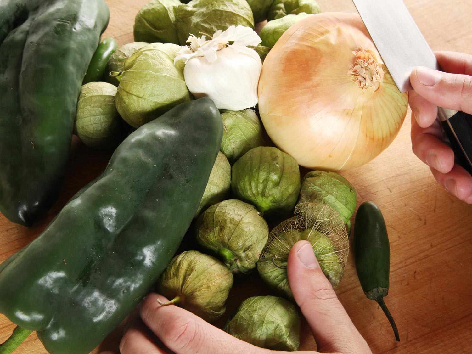 A pile of unprepared ingredients for the green sauce: Poblanos, tomatillos, garlic, onion, and serranos. A hand holding a kitchen knife is poised to cut..