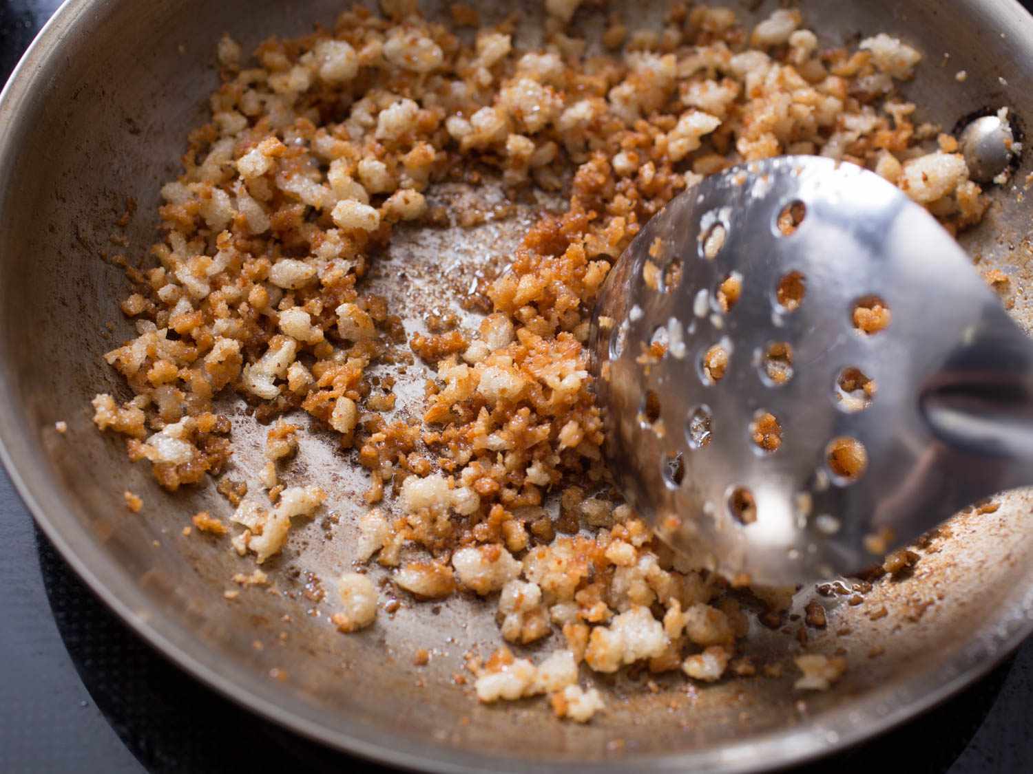 A slotted spoon stirs small, browned pieces of bacon on a skillet. 