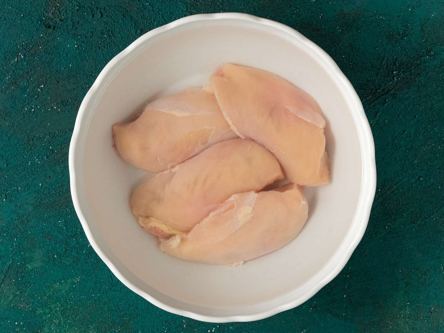 Chicken breast halves submerged in brine inside of a white ceramic bowl on a teal background.