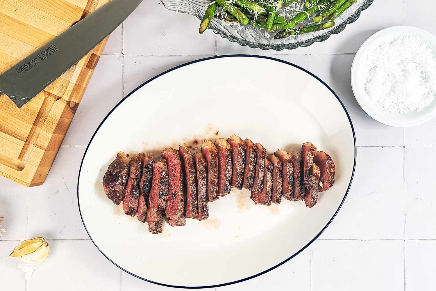 Sliced cooked steak on a white plate kitchen tools and side dishes visible nearby