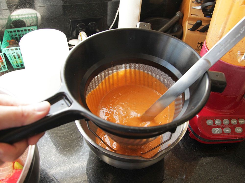 Pressing blended gazpacho through a fine-mesh strainer with a ladle. 