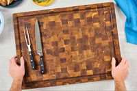 An overhead view of a Yes4All Teak Cutting Board with a meat fork and slicing knife on the lefthand side