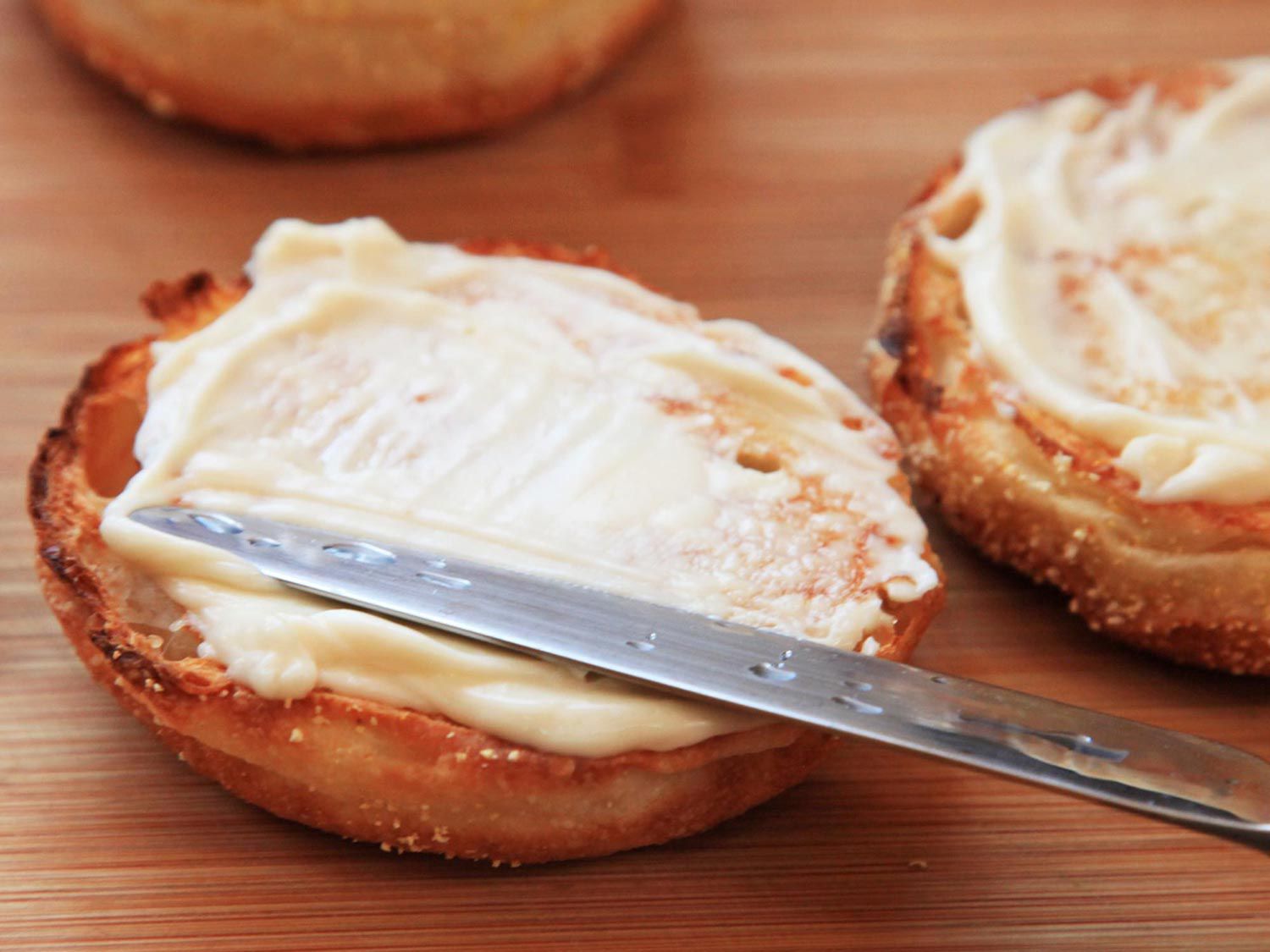 A butter knife spreading mayo on a toasted English muffin