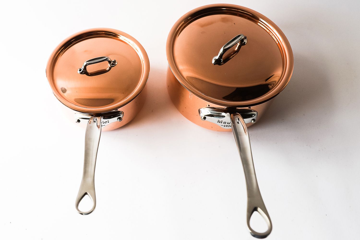 Two copper saucepans with lids featuring silver handles placed side by side on a white surface