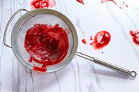 Rösle Stainless Steel Round Handle Kitchen Strainer with redcolored puree pressed inside placed on a countertop with smeared remnants around it